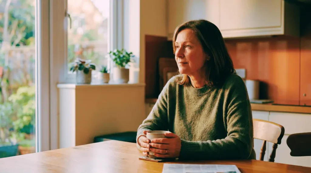 woman sitting at table in kitchen holding a mug and considering whether to hire a caregiver